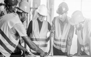 Four people in hard hats and hi-vis vests reading drawings on site, in black and white