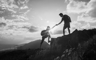 One climber reaching down to pull another onto a ridge at sunrise, in black and white
