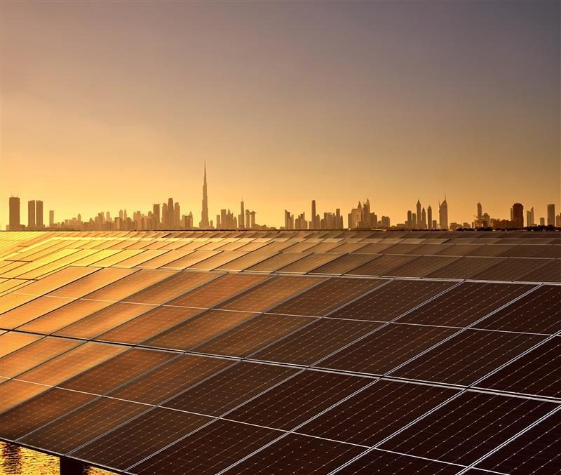 A rooftop array of solar panels at sunset, with the Dubai skyline and the Burj Khalifa on the horizon.
